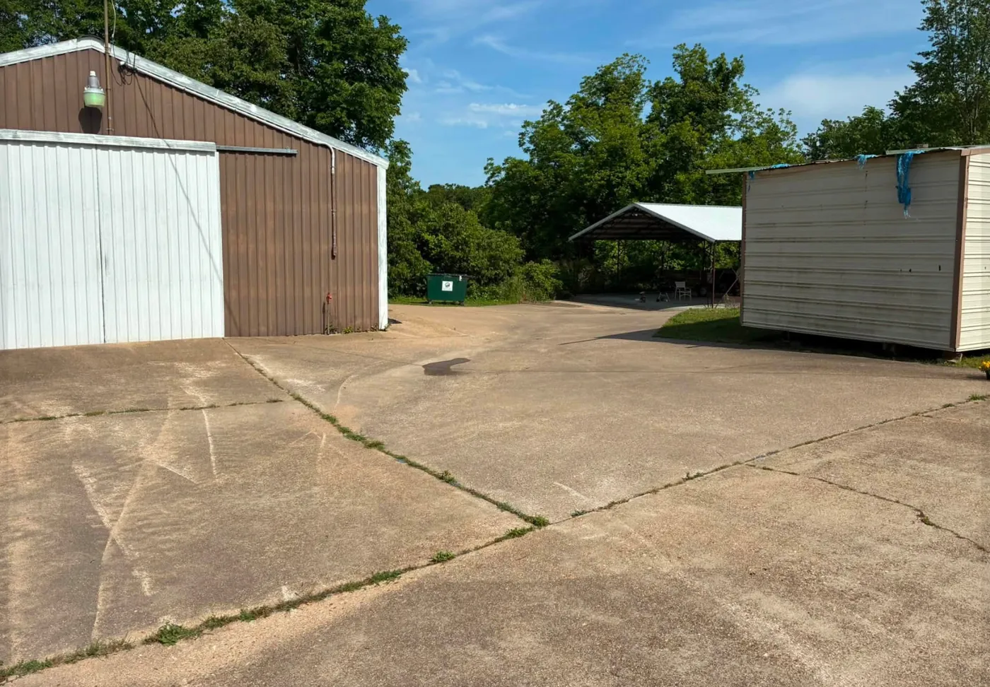Concrete lot in front of a metal building and shed, gray and dirty, with weeds along the cracks