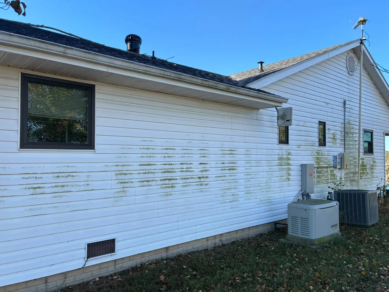 White vinyl siding streaked with green algae beside a generator and an air conditioner