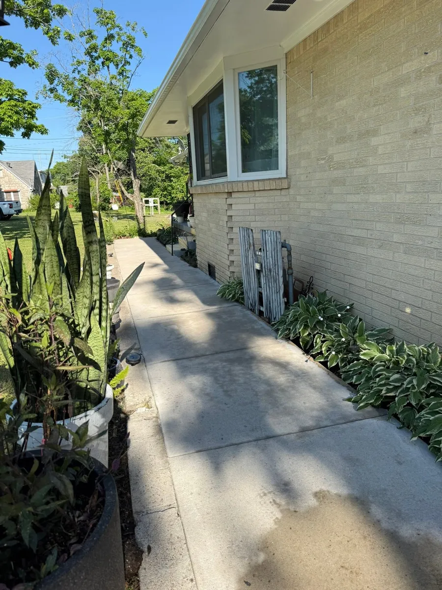 Concrete sidewalk running beside a yellow brick house with garden beds